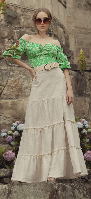 Mujer vistiendo maxi falda color beige, blusa manga 3/4 verde, correa beige trenzada y gafas ovaladas color café
