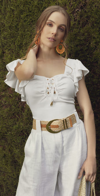 Modelo vistiendo blusa tipo crop blanca, pantalón blanco, cinturón color beige y aretes color naranja de la marca Studio F